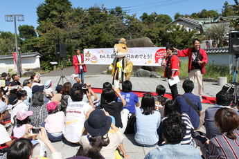 お祝いのメッセージが書かれた横断幕の前でキラキラ衣装のわん丸君