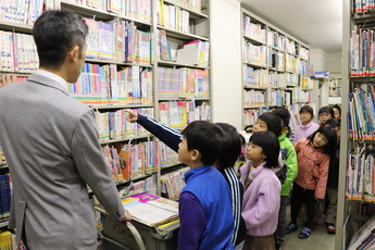 図書館職員の男性を先頭に本棚の前で説明を受ける児童たち