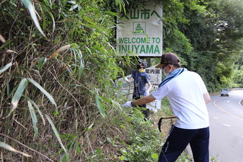 道路脇で看板を覆っている草をカマで刈る男子学生