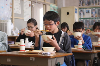 教室の前向きの机で給食を食べるメガネをかけた男児