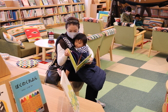 図書に囲まれた空間で赤ちゃんを抱っこした女性が絵本を手にとる様子